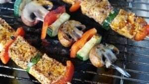 Skewers with seasoned vegetables and tofu, including mushrooms, zucchini, and bell peppers, grilling on a barbecue grill beside savory meat skewers.