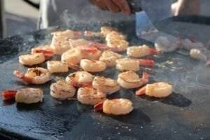 Person grilling seasoned shrimp on a large flat-top grill with a spatula, surrounded by the serene sounds of nature. Steam rises from the cooking shrimp.
