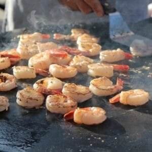 Close-up of a person BBQ cooking shrimp on a flat-top grill. The shrimp are sizzling and slightly charred, with visible steam rising.