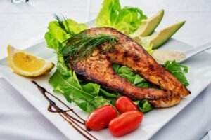 Grilled salmon steak on a bed of leafy greens, garnished with dill, accompanied by tomato and avocado slices, and a lemon wedge on a white plate with a drizzle of sauce.