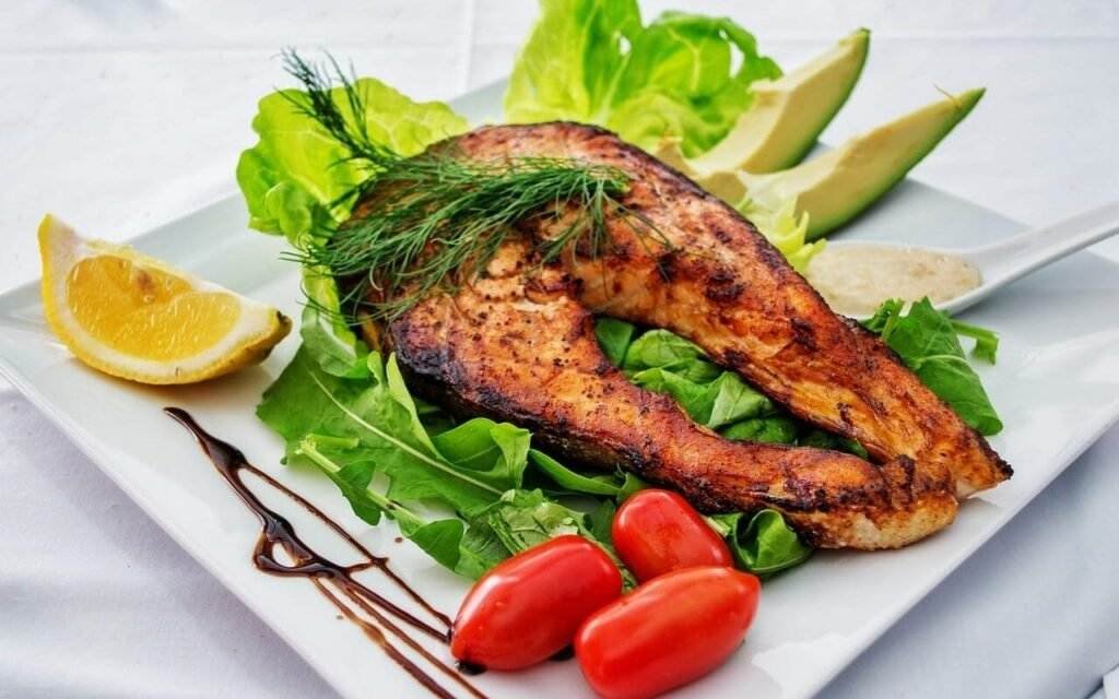 Grilled salmon steak served on a bed of lettuce with cherry tomatoes, avocado slices, a lemon wedge, and a drizzling of balsamic vinegar on a white square plate.