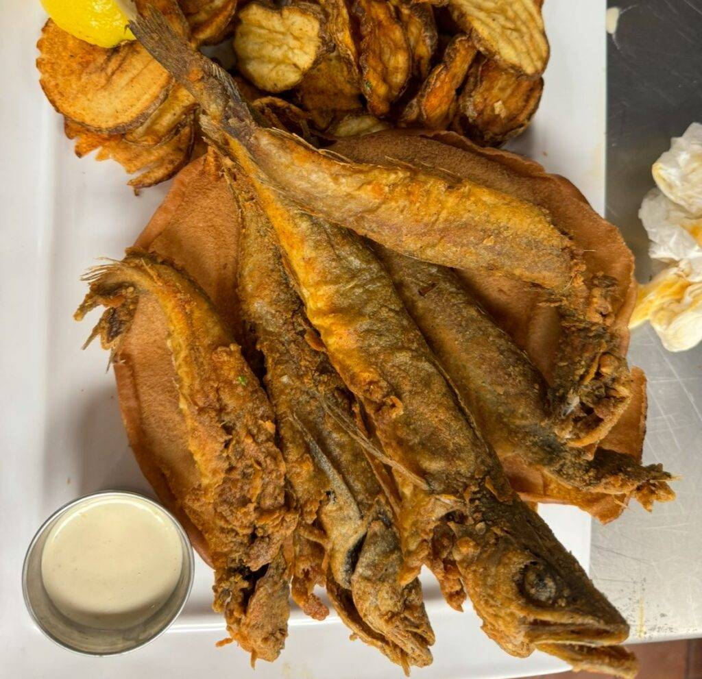 A plate with several pieces of fried whole fish, a serving of sliced fried potatoes, a lemon wedge, a small cup of dipping sauce, and a crumpled napkin on the side. A plate with several pieces of fried whole fish, a serving of sliced fried potatoes, a lemon wedge, a small cup of dipping sauce, and a crumpled napkin on the side.