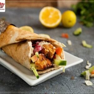 A chicken shawarma wrap with vegetables is served on a rectangular white plate, resembling a Chicken Sandwich (ساندوتش دجاج), with lemons and scattered salad pieces in the background.